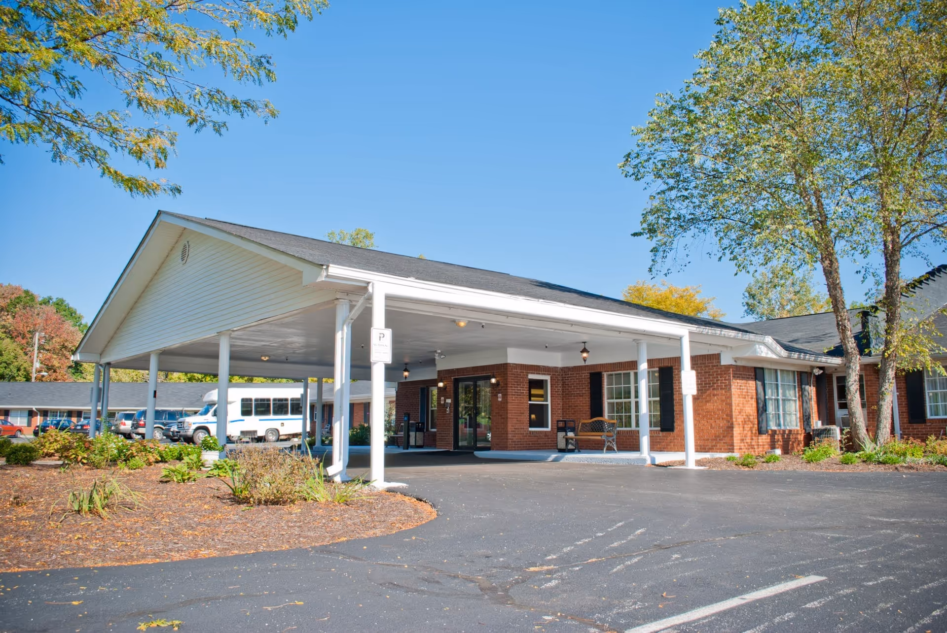 Exterior view of Harcourt Terrace Nursing and Rehabilitation facility showing a covered entrance with white pillars, a brick building, a bench near the entrance, parked vehicles including a white shuttle bus, and trees with green and autumn-colored leaves under a clear blue sky.