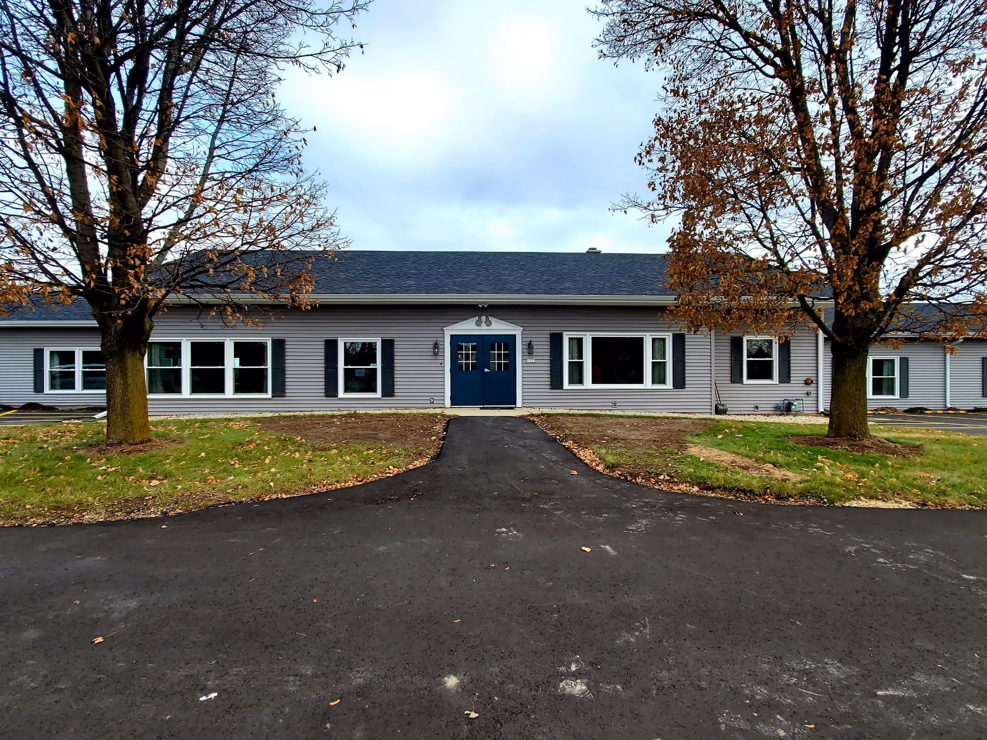 Front exterior view of a single-story building with gray siding, a dark roof, and blue double doors in the center. There are multiple windows with black shutters on either side of the entrance. Two trees with sparse brown leaves flank the walkway leading to the entrance. The sky is overcast.
