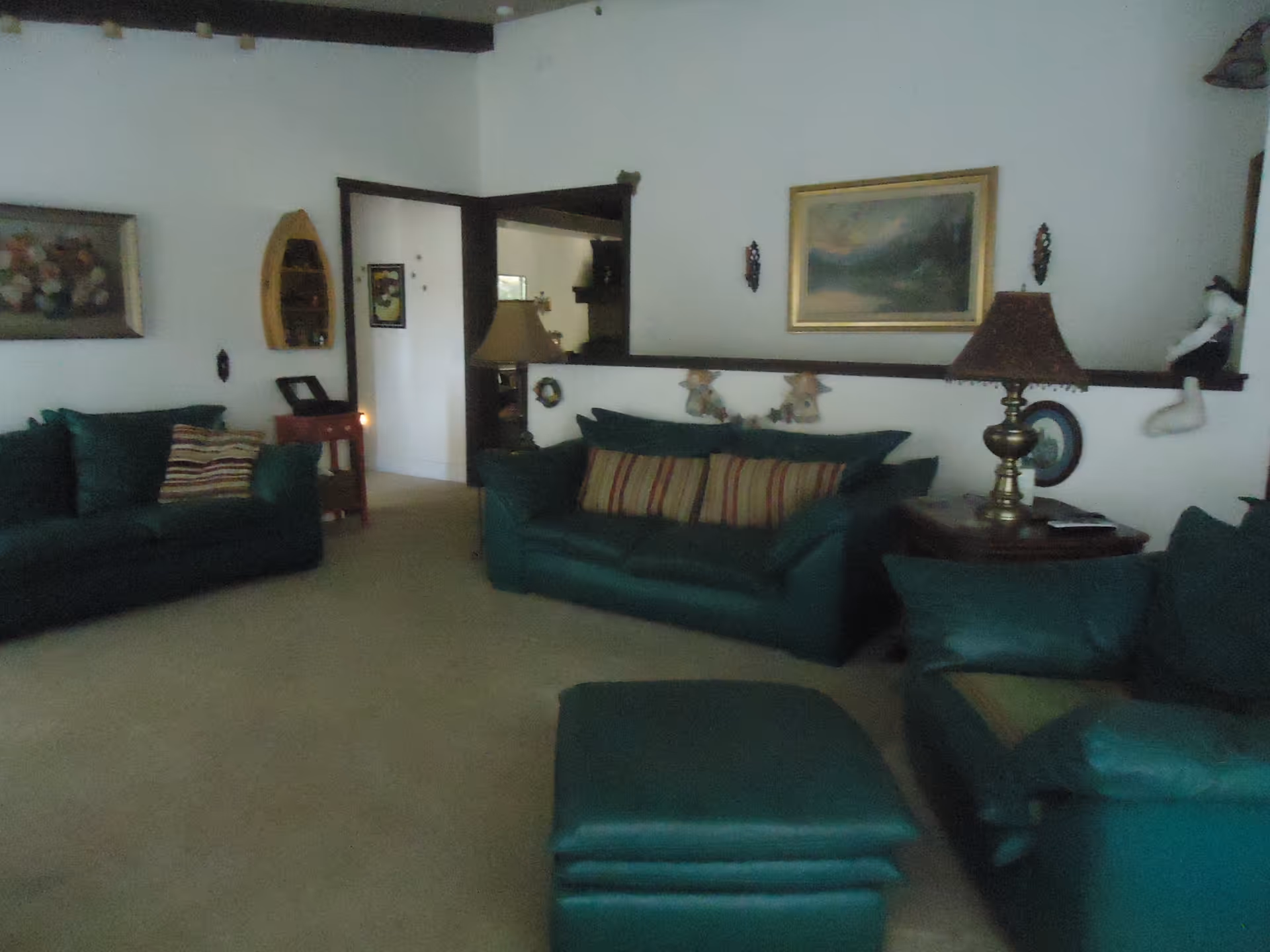 A living room with green upholstered sofas and a matching ottoman. There are striped pillows on the sofas, a wooden side table with a brass lamp, and framed artwork on the white walls. The room has carpeted flooring and visible dark wooden beams on the ceiling.