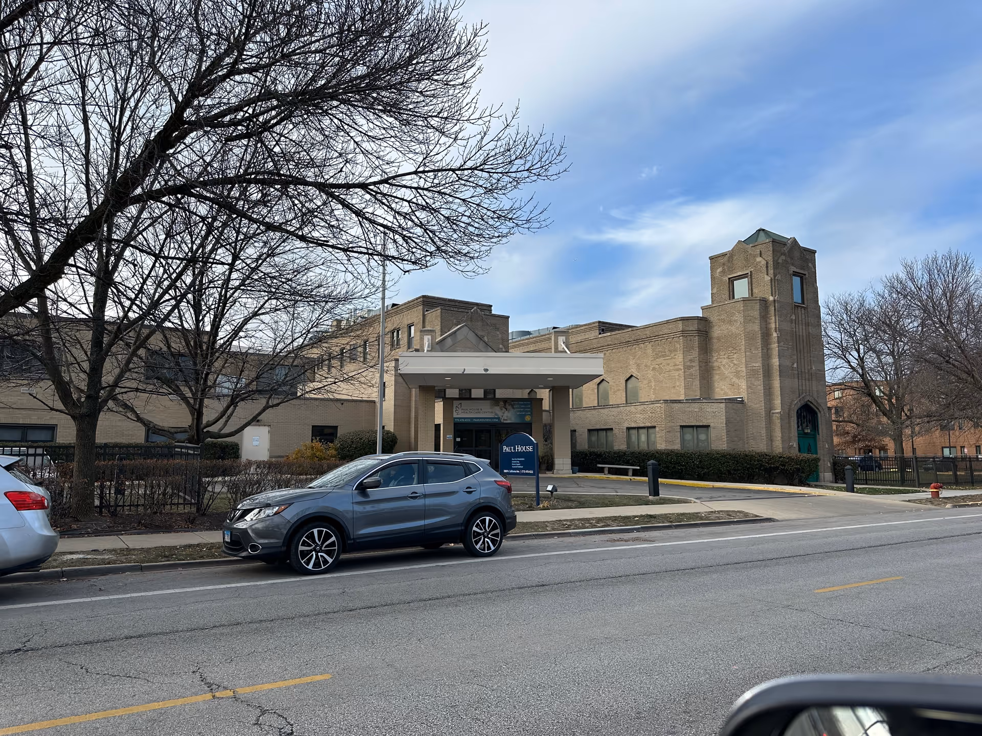 Exterior view of Paul House and Healthcare Center, a large beige brick building with a covered entrance and a tower-like structure on the right side. There are leafless trees and parked cars along the street in front of the building under a partly cloudy sky.
