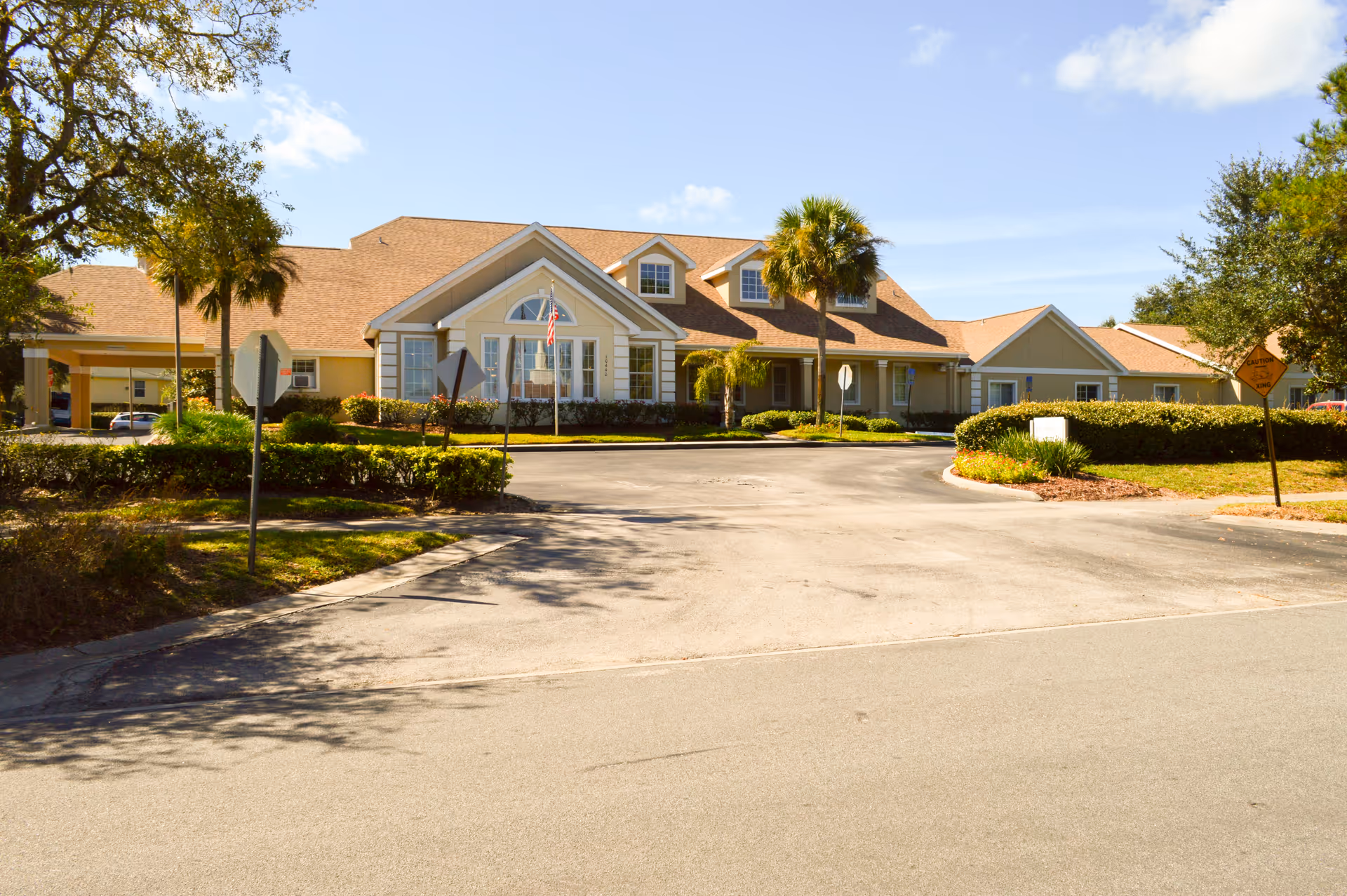 Exterior view of a single-story senior living facility building with beige walls and a brown shingle roof under a clear blue sky. The building has multiple windows, a covered entrance, and is surrounded by palm trees, shrubs, and a paved driveway.
