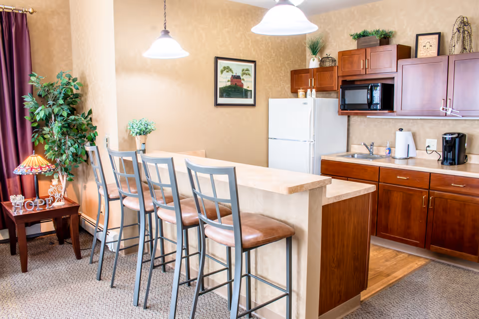 A bright kitchen area in a senior living facility featuring a countertop with four high chairs, wooden cabinets, a white refrigerator, a microwave, a sink, and a coffee maker. There is a small table with a decorative lamp and a plant next to a window with purple curtains. The walls are beige with a framed picture hanging above the refrigerator.