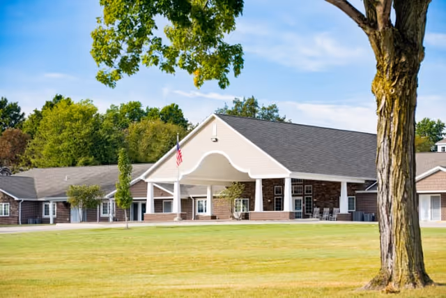 Exterior view of a single-story senior living facility building with a covered entrance, an American flag, and a large tree in the foreground on a sunny day with a blue sky and scattered clouds.