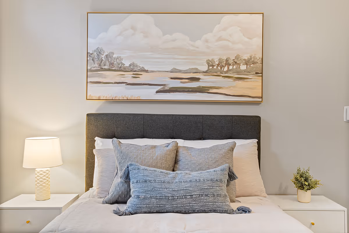 A neatly made bed with a dark gray upholstered headboard, adorned with multiple pillows in shades of gray and blue. On either side of the bed are white nightstands; the left nightstand has a white textured lamp with a light shade, and the right nightstand has a small potted plant. Above the bed hangs a large framed painting depicting a serene landscape with clouds, trees, and water in soft neutral tones.