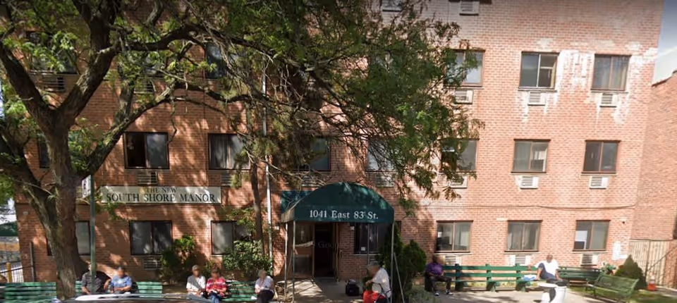 Exterior view of The New South Shore Manor, a brick multi-story building with several windows. There is a green awning above the entrance displaying the address 1041 East 83 St. Several people are sitting on green benches in front of the building under the shade of a large tree.