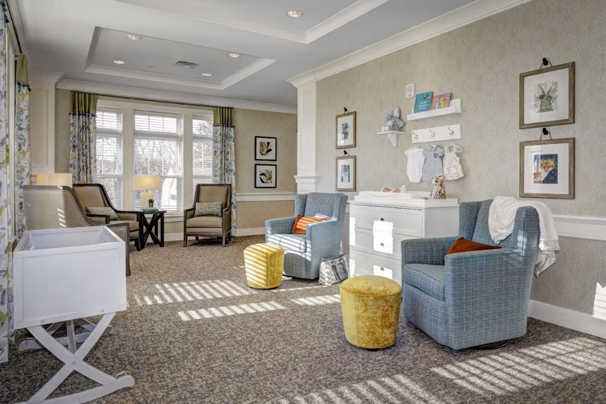 A bright and cozy sitting area in a senior living facility with two blue armchairs, two yellow ottomans, a white dresser with baby clothes and toys displayed above it, and a white bassinet. The room has large windows with floral curtains, framed artwork on the walls, and carpeted flooring.