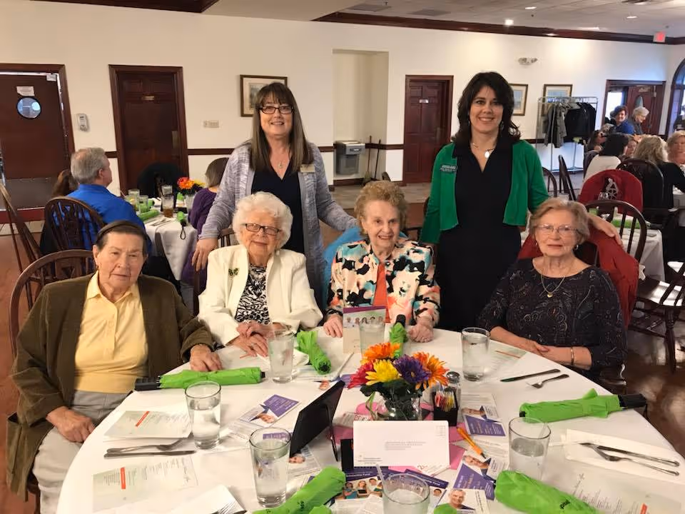 A group of elderly women seated around a dining table with two standing women behind them in a well-lit room. The table is set with menus, glasses of water, green napkins, and a colorful flower centerpiece. Other people are visible in the background seated at tables.