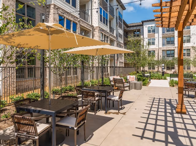 Outdoor patio area at Album Keller Ranch with tables and chairs under large beige umbrellas, surrounded by greenery and a multi-story residential building in the background under a clear blue sky.