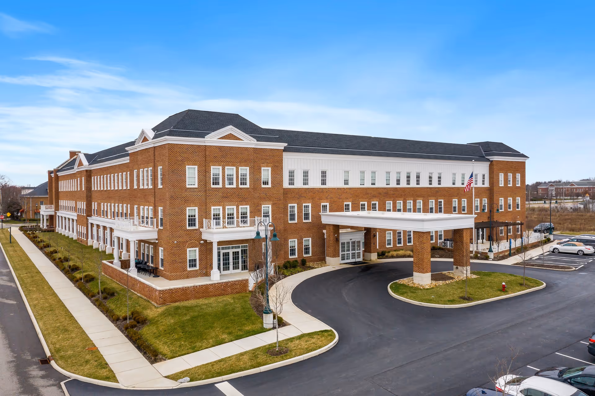 Exterior view of a large three-story brick building with white trim and multiple windows, featuring a covered entrance with columns and an American flag. The building is surrounded by a paved driveway, parking spaces, sidewalks, and landscaped grass areas under a blue sky.
