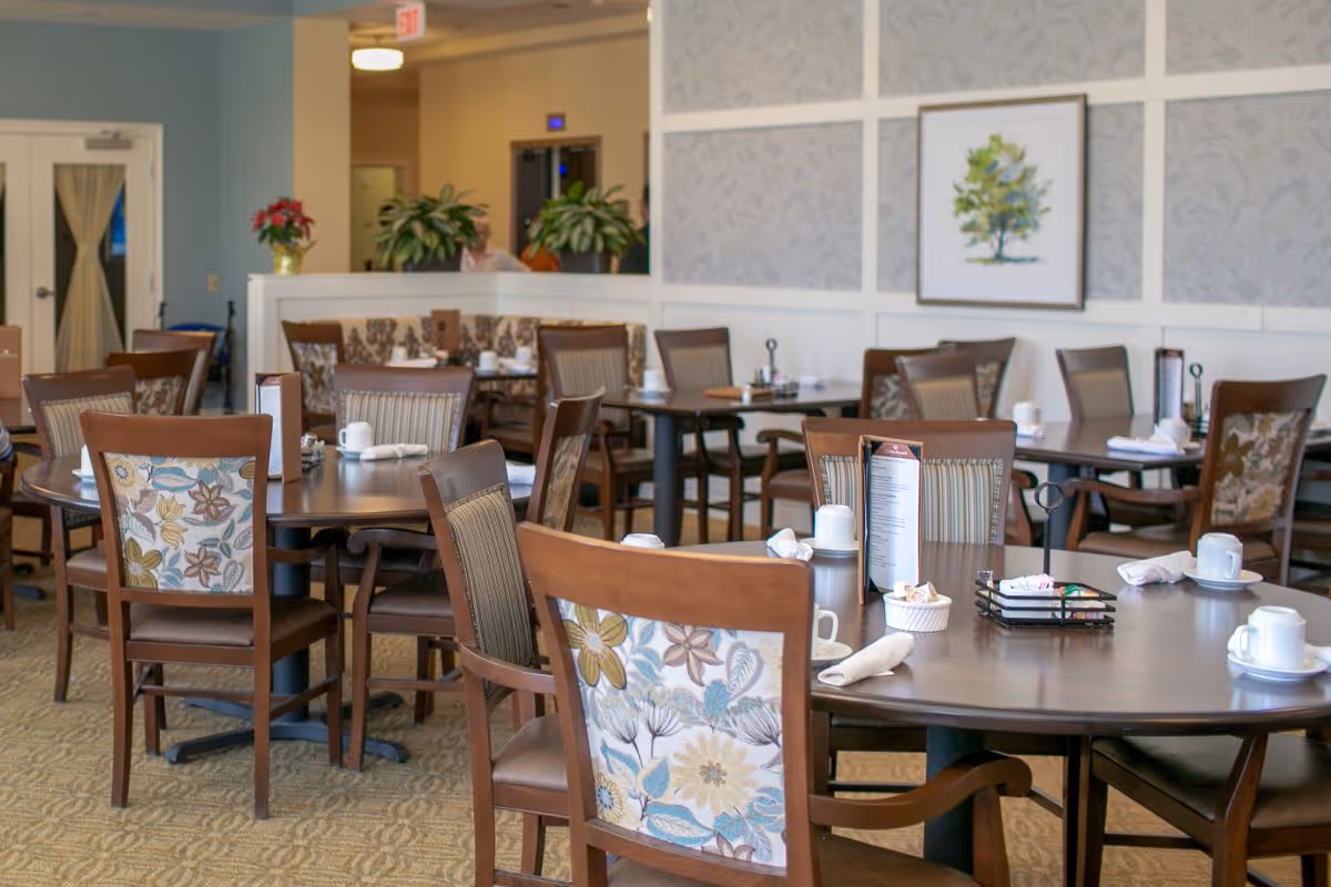 A dining room in a senior living facility with multiple wooden tables and chairs. The chairs have floral and striped upholstery. Tables are set with white cups, napkins, and menus. The room has light-colored walls with decorative paneling and a framed picture of a tree. There are plants and a door visible in the background.
