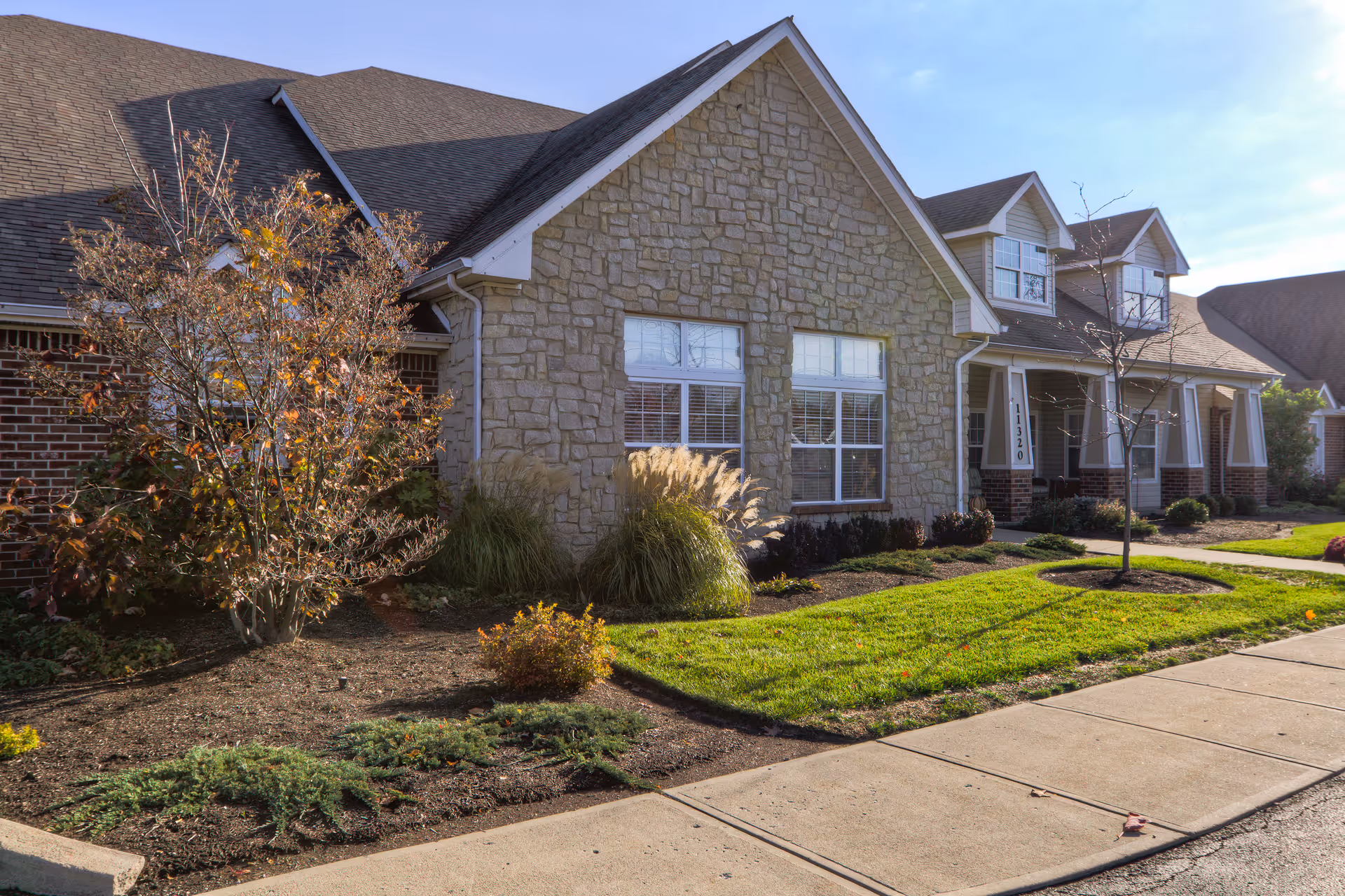 Exterior view of a senior living facility building with stone and brick facade, multiple windows, a porch with columns, and landscaped greenery including bushes and a small tree along a sidewalk.