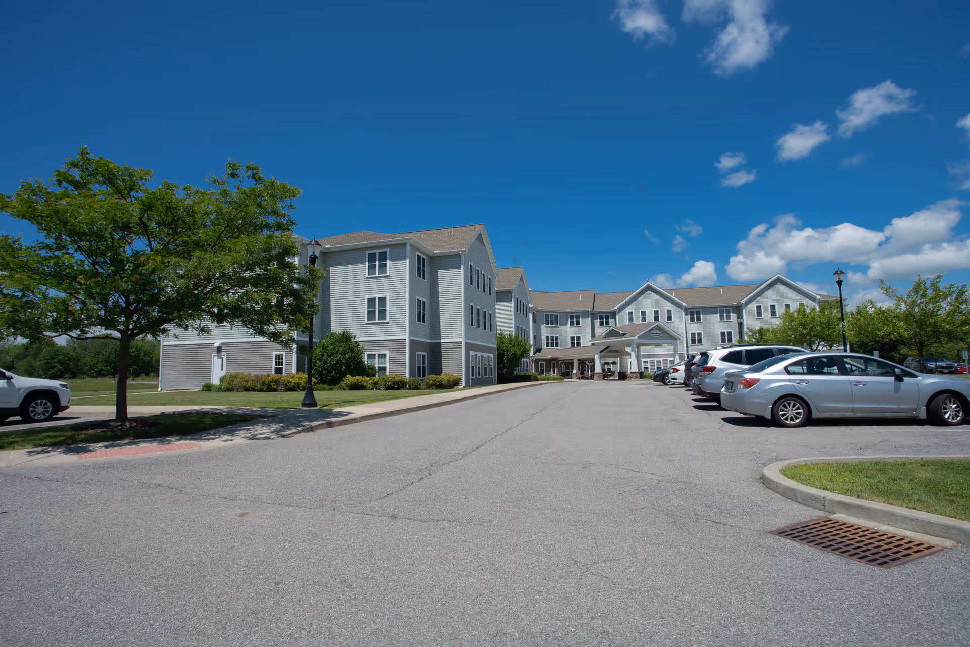 Exterior view of Judson Meadows Assisted Living facility on a sunny day with a clear blue sky. The building is a large, multi-story structure with light gray siding and white trim. There is a parking lot in front with several parked cars, a tree on the left side, and a sidewalk leading to the entrance.