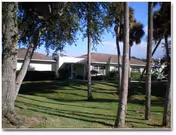 Single-story residential building with a brown roof surrounded by green grass and several tall trees under a clear blue sky.
