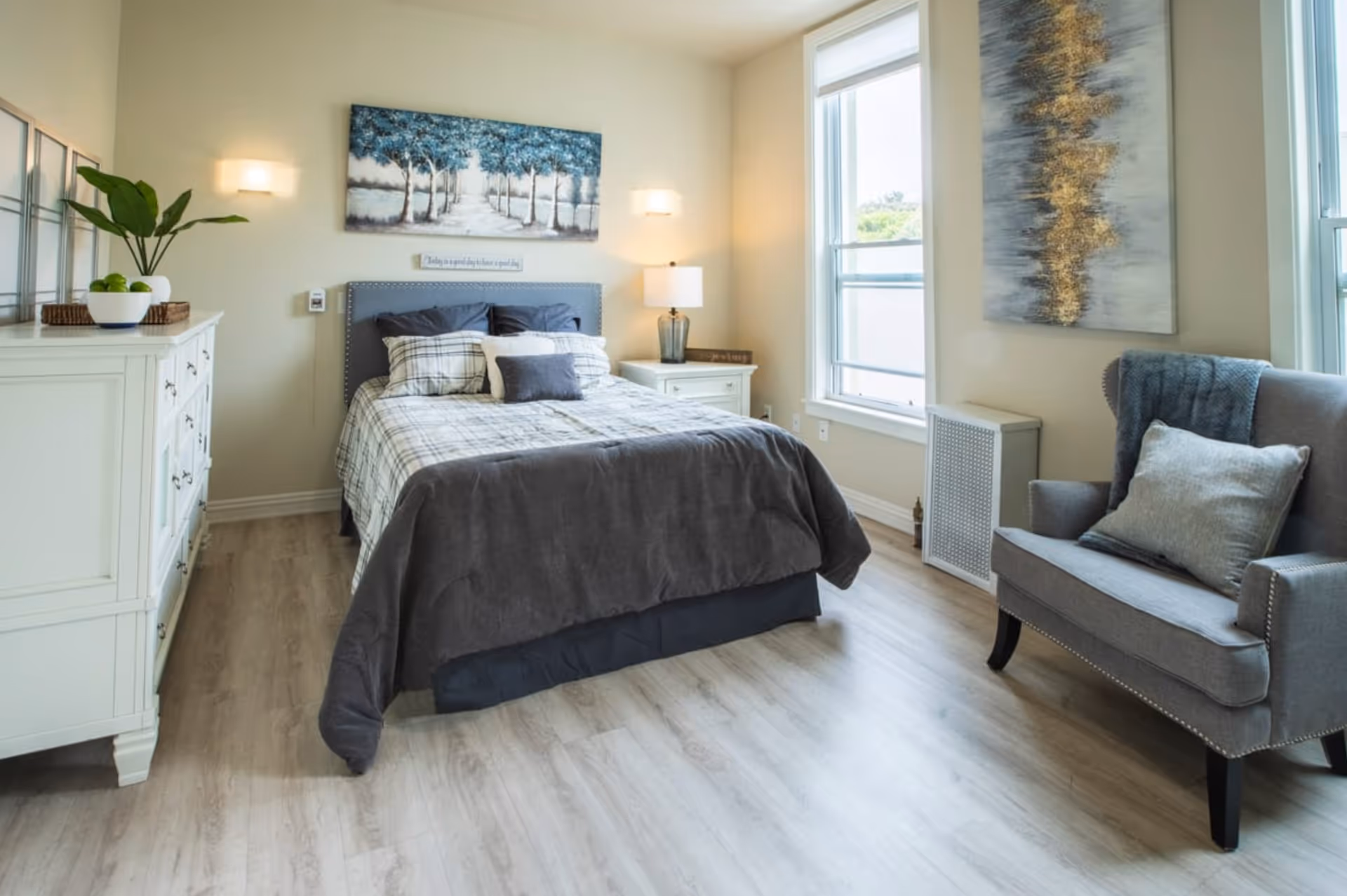 A bright and cozy bedroom with a bed featuring plaid bedding and multiple pillows, a white dresser with a plant and bowl of green apples, a nightstand with a lamp, two large windows letting in natural light, a gray armchair with a pillow and throw blanket, and two pieces of wall art above the bed and chair.