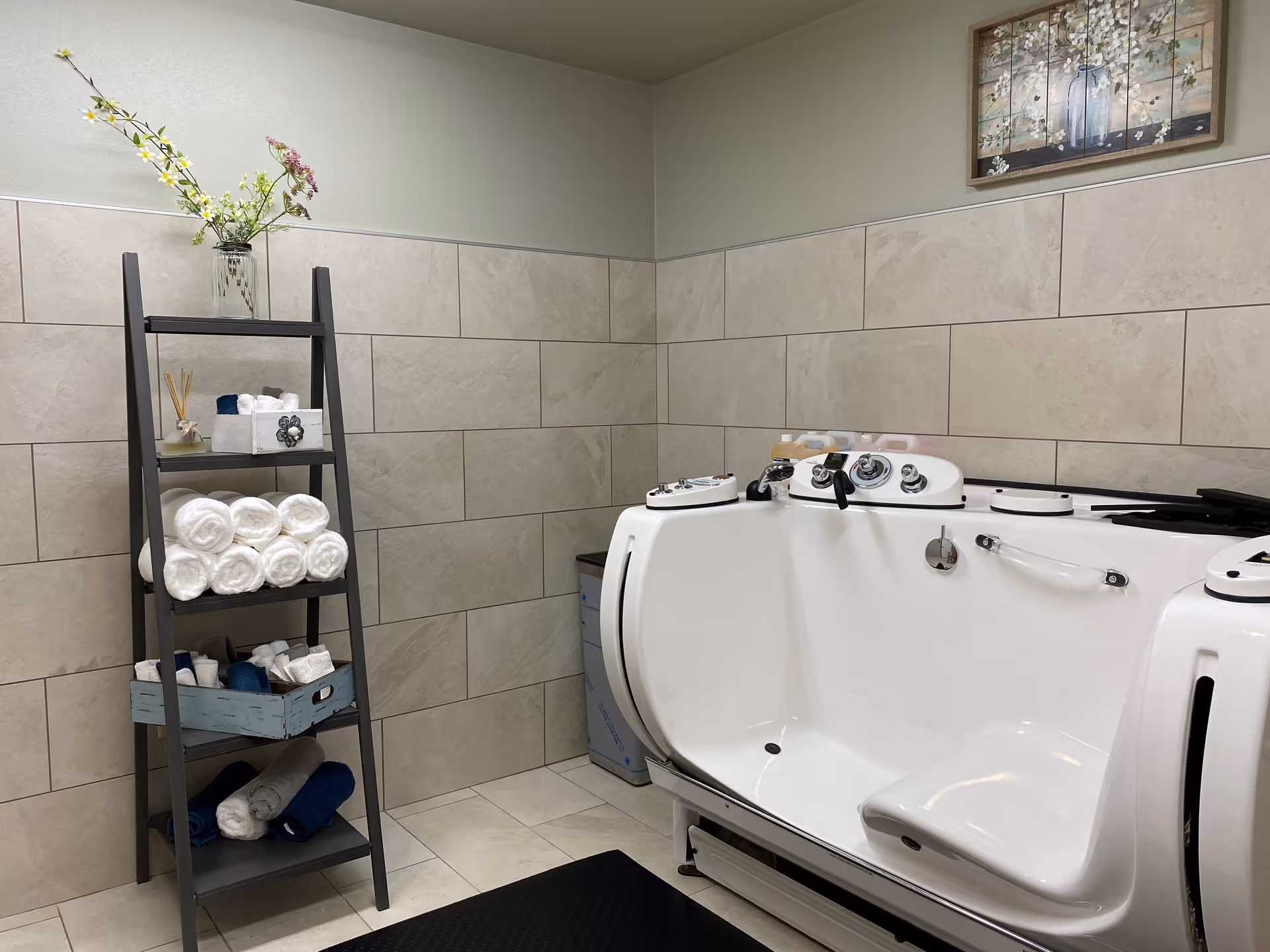 A bathroom with beige tiled walls and floor featuring a white walk-in bathtub with multiple controls and a seat. To the left, there is a dark gray ladder-style shelf holding rolled white and blue towels, a small basket, and a glass vase with flowers. A framed floral painting hangs on the wall above the bathtub.