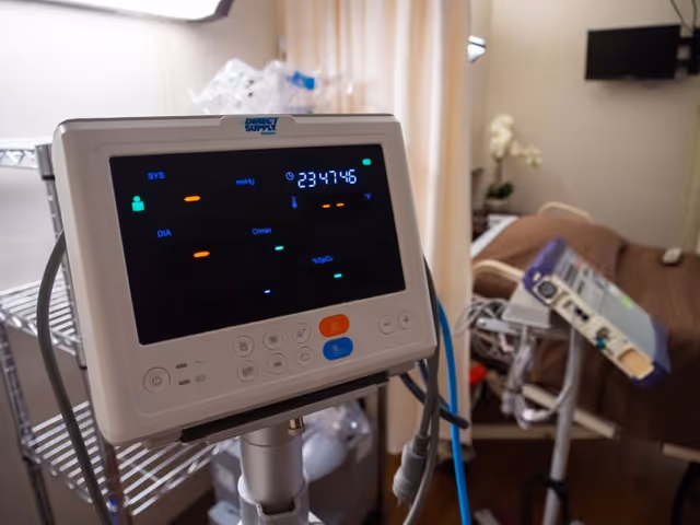 Close-up of a medical monitor displaying vital signs in a patient room with a hospital bed and medical equipment in the background.