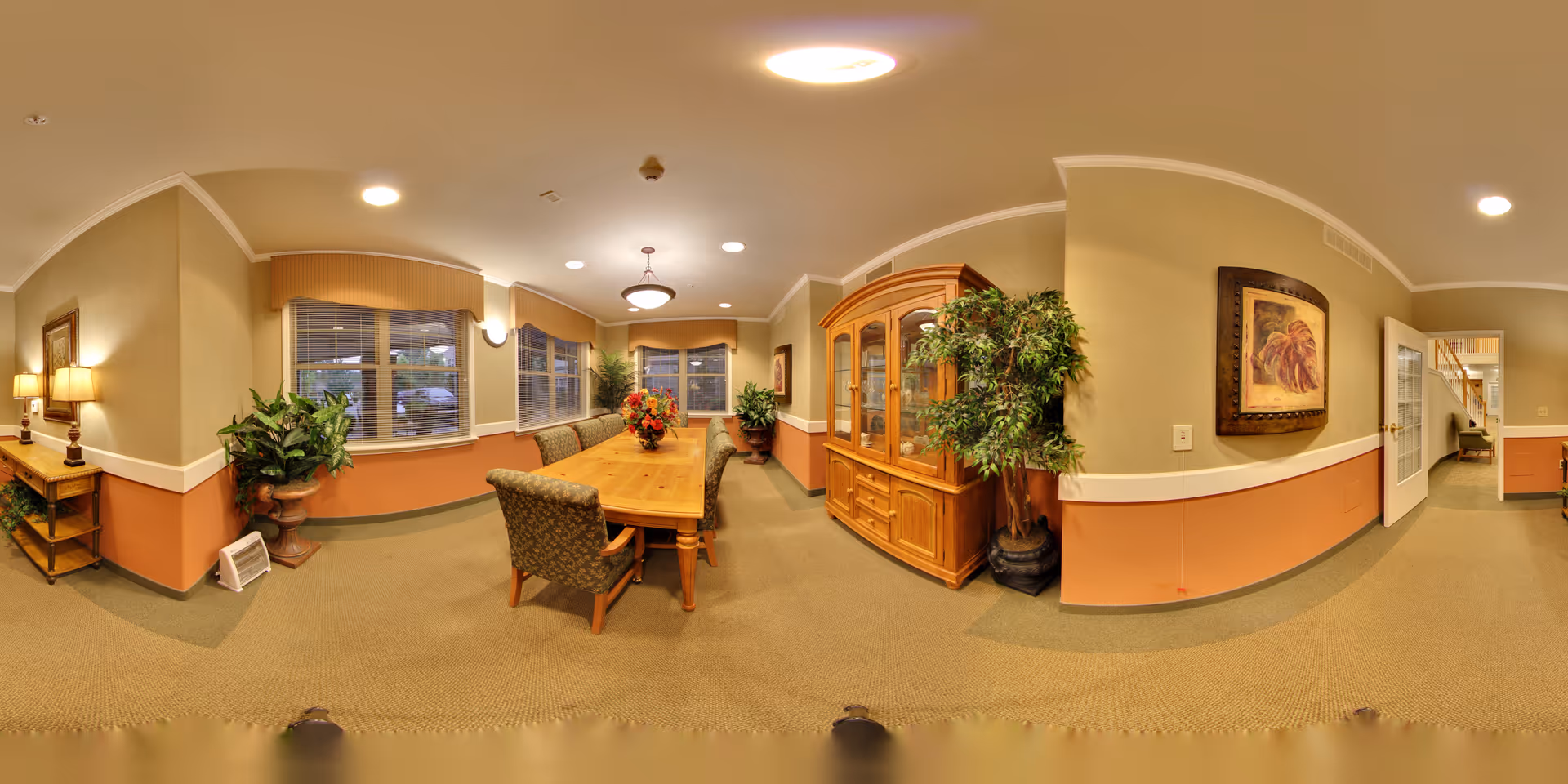 A warmly lit dining room in a senior living facility with a long wooden table surrounded by upholstered chairs. The room features large windows with valances, a wooden china cabinet, potted plants, framed artwork on the walls, and a side table with lamps. The carpet is green and the walls are painted beige with a darker orange accent on the lower half.