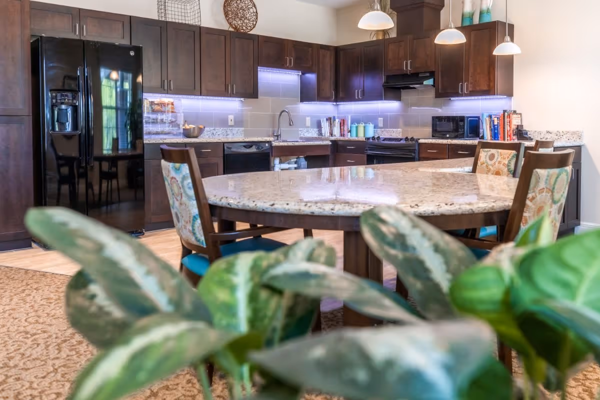 A modern kitchen with dark wood cabinets, a black refrigerator, a stove, a microwave, and a dishwasher. There is a large round granite table surrounded by chairs with colorful patterned upholstery. Green plants are visible in the foreground.