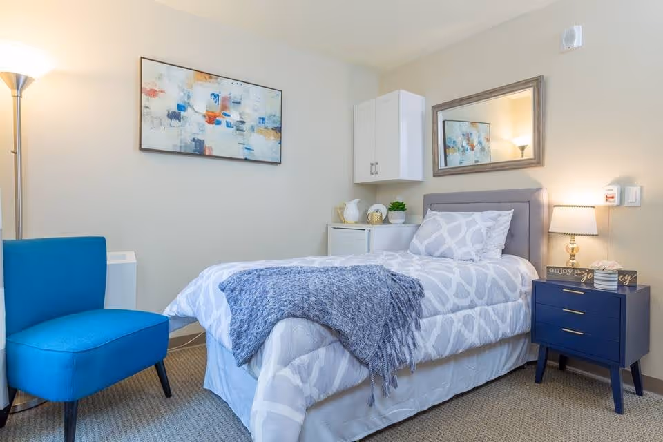 A neatly made single bed with white and gray patterned bedding and a blue throw blanket draped over the foot. Next to the bed is a navy blue nightstand with a lamp, a small plant, and decorative items. A large mirror hangs on the wall above the bed. To the left of the bed is a bright blue upholstered chair and a tall floor lamp. The walls are light-colored and there is abstract artwork hanging above the chair.