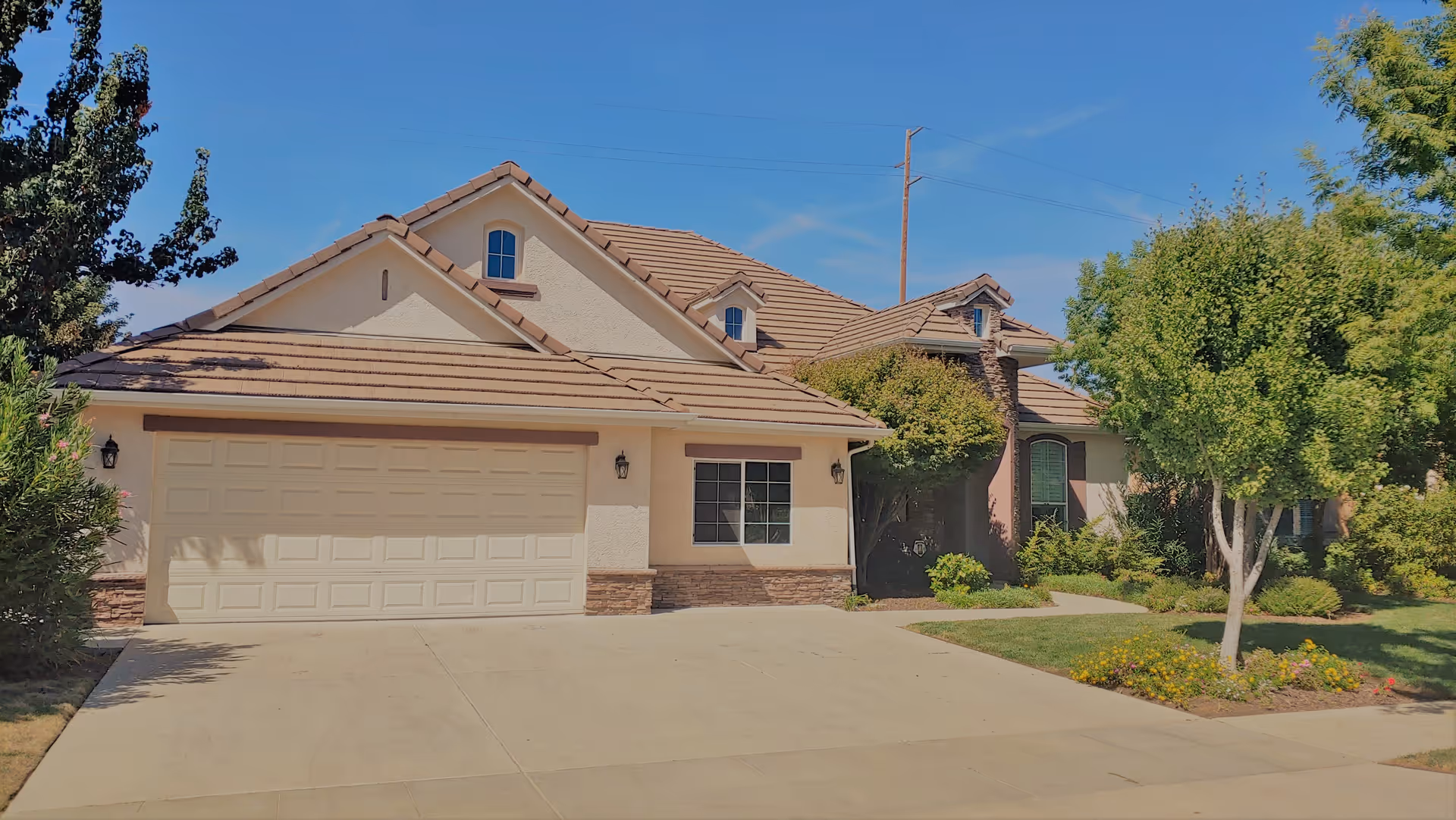 Front exterior view of a single-story residential home with a two-car garage, beige stucco walls, brown tiled roof, and a well-maintained front yard with trees and shrubs under a clear blue sky.