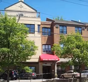 Exterior view of a multi-story brick and stone building with a red awning over the entrance. There are several windows and green trees in front of the building, with parked cars along the street.