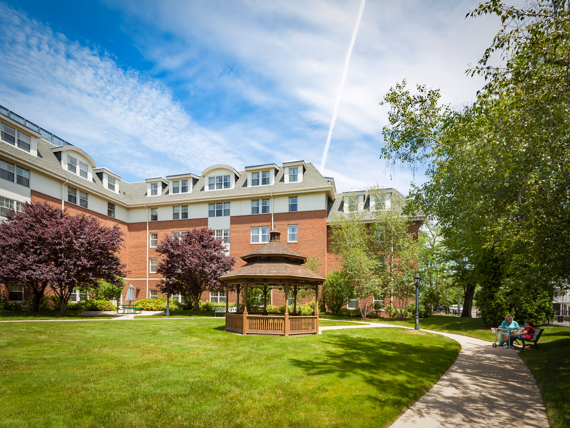 A sunny outdoor garden area at Providence House Assisted Living featuring a wooden gazebo in the center of a green lawn, surrounded by trees and shrubs. A paved walkway curves through the garden, where two people are sitting on a bench near the right side of the image. The background shows a multi-story brick building with many windows under a blue sky with some clouds.