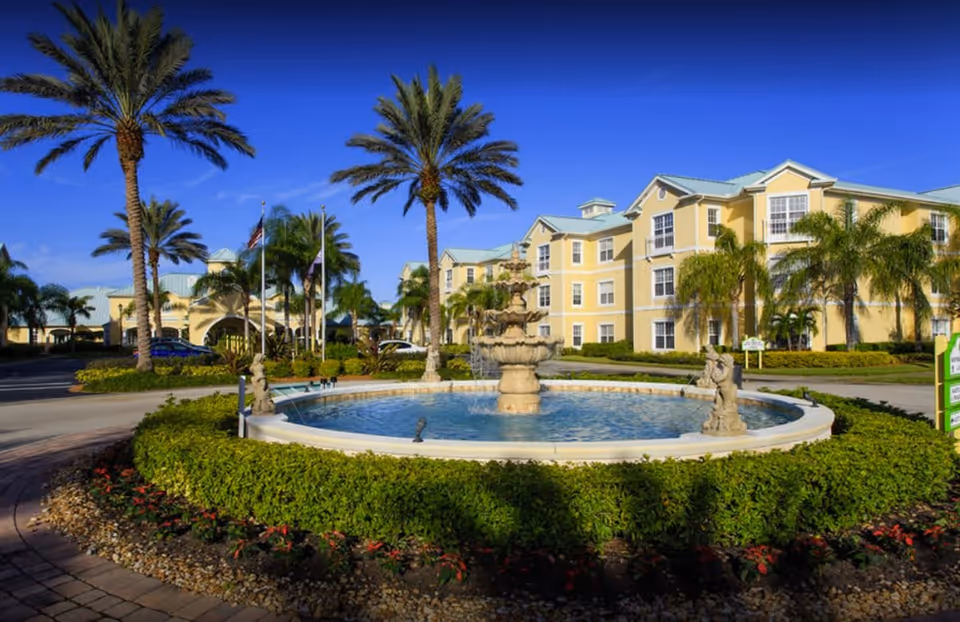 Exterior view of The Brennity at Vero Beach Senior Living facility featuring a large circular fountain surrounded by greenery and palm trees, with a yellow multi-story building in the background under a clear blue sky.