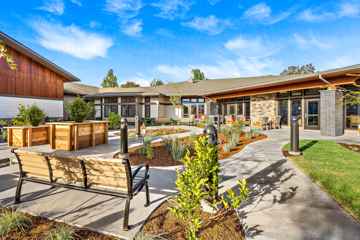 Outdoor courtyard area of the Memory Support Center at Rogue Valley Manor featuring a paved walkway, wooden benches, raised garden beds, various plants, and a building with large windows and a covered patio with seating under a bright blue sky with some clouds.