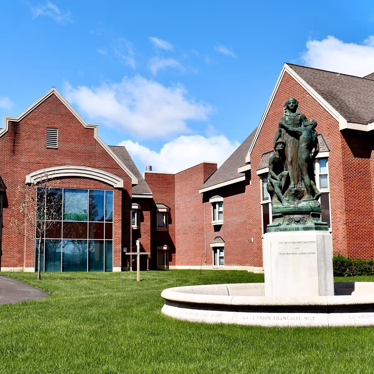 Exterior view of a red brick building with large windows and a statue in front on a green lawn under a blue sky with some clouds.