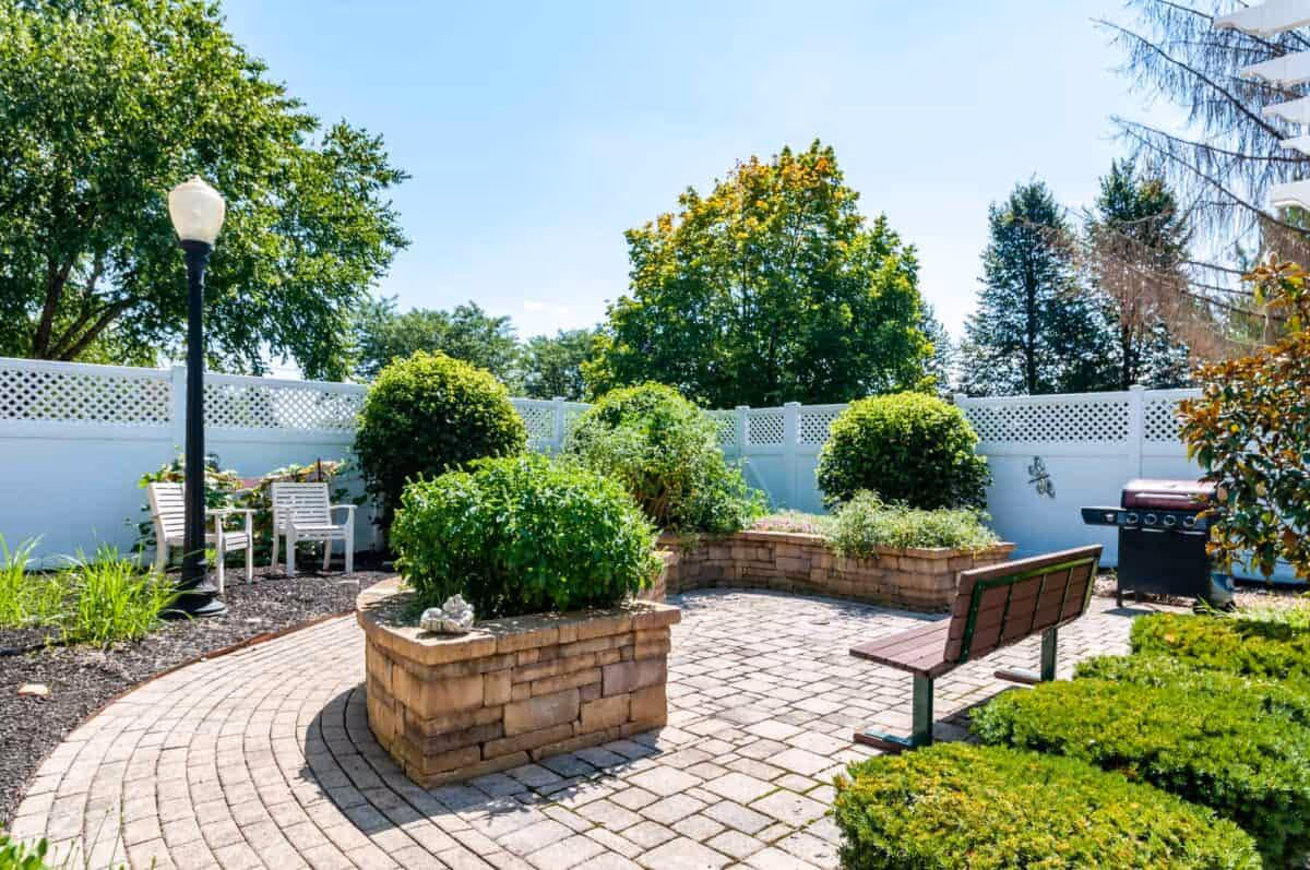 A sunny outdoor garden area with a curved brick planter filled with green shrubs and plants. There is a wooden bench with a metal frame facing the planter, a black streetlamp, two white chairs, and a barbecue grill near a white lattice fence. Trees with green foliage are visible in the background under a clear blue sky.