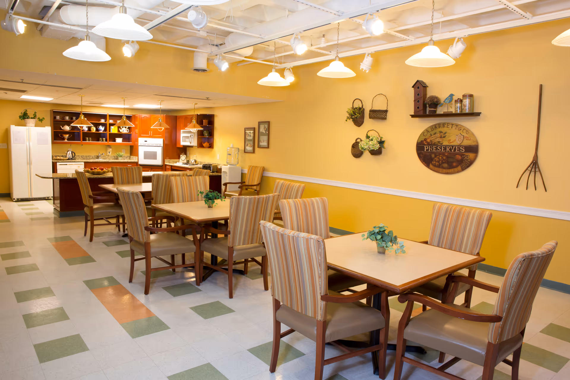 A warm and inviting dining area with several square tables surrounded by striped cushioned chairs. The room features a yellow wall decorated with hanging baskets, a birdhouse, and a round wooden sign that says 'Preserves.' In the background, there is a kitchen area with wooden cabinets, a white refrigerator, oven, microwave, and a water cooler. The floor has a patterned tile design in green, white, and orange, and the ceiling has exposed pipes and multiple hanging light fixtures.