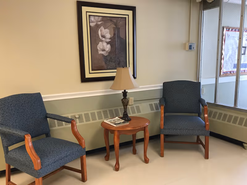 A small seating area with two blue upholstered armchairs with wooden armrests, a wooden side table between them holding a decorative lamp and a magazine. A framed floral artwork hangs on the beige wall behind the chairs. There is a window with a wire mesh on the right side and a thermostat on the wall.