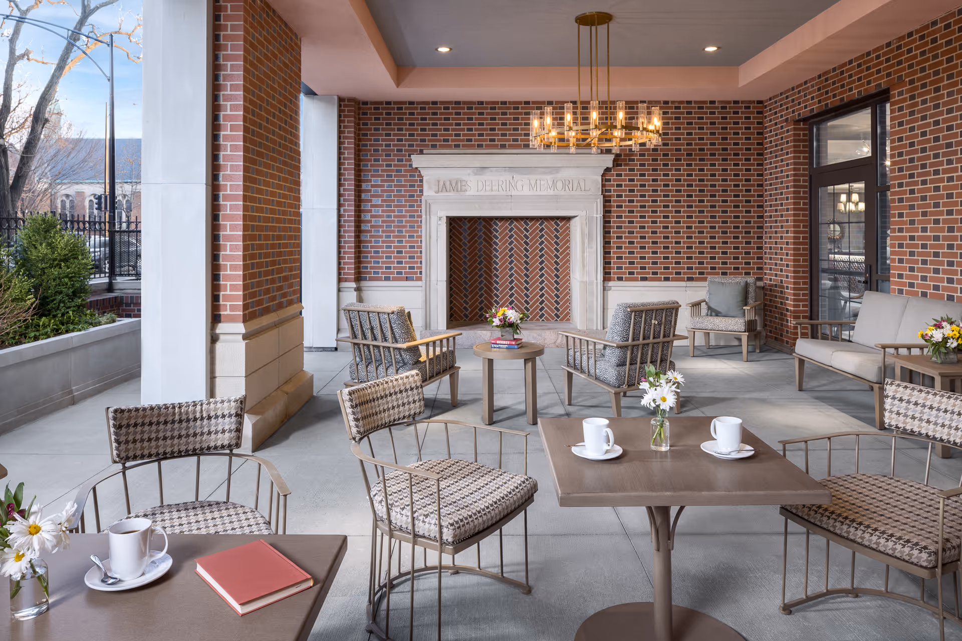 Outdoor covered patio area with brick walls and a decorative fireplace labeled 'James Deering Memorial'. The space is furnished with metal chairs featuring patterned cushions, small tables with flowers and coffee cups, and a chandelier hanging from the ceiling. There are plants and a fence visible outside the patio.
