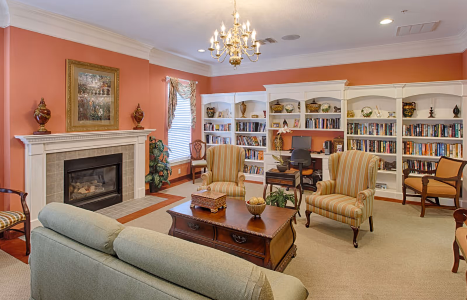 A cozy senior living room with a green sofa, two striped armchairs, a wooden coffee table, and a fireplace with decorative vases and a framed painting above it. There is a large white bookshelf filled with books and decorative items, and a small desk with a computer and chair. The walls are painted a warm orange color, and a chandelier hangs from the ceiling.