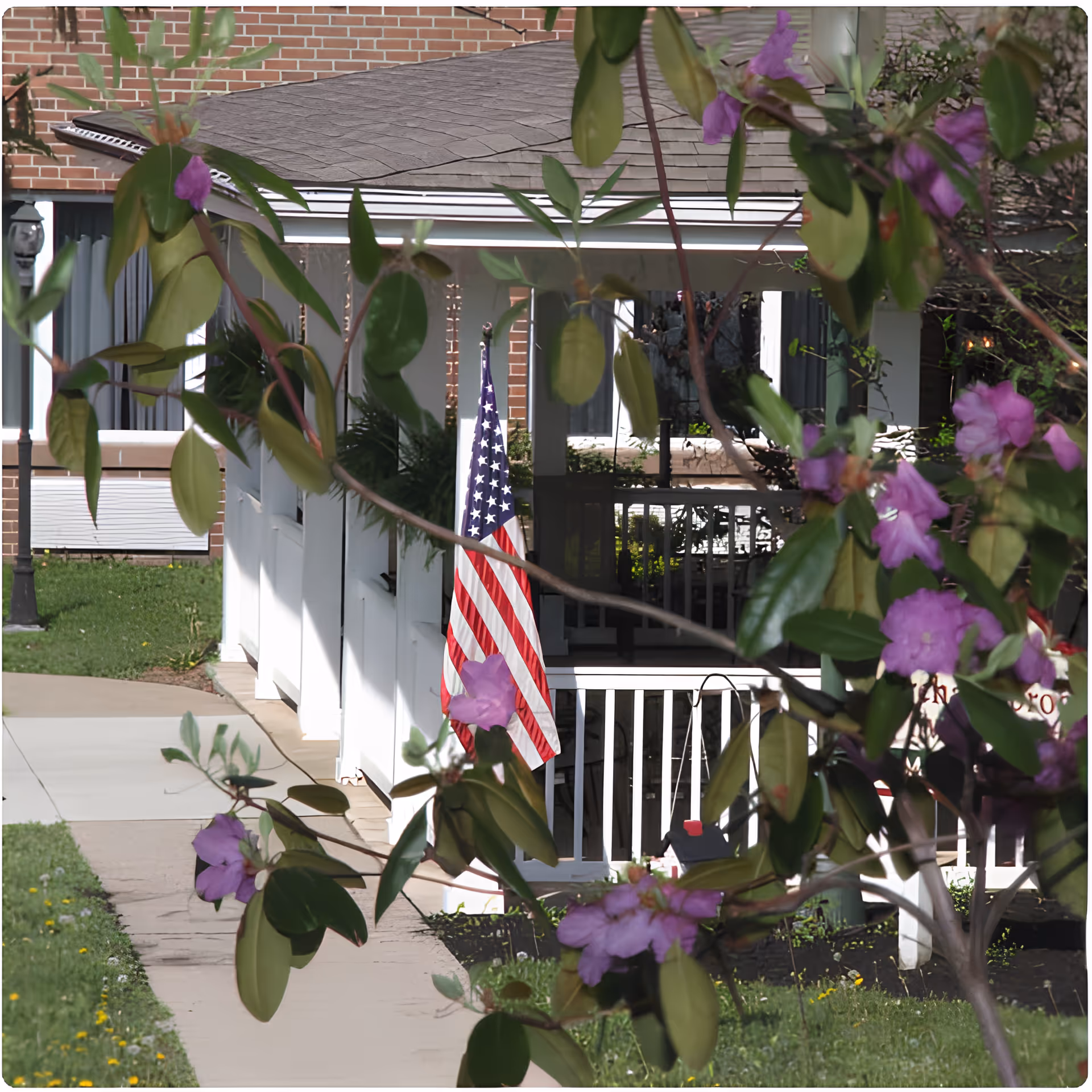 View of a white porch with an American flag hanging, partially obscured by branches with green leaves and purple flowers. The porch is attached to a brick building with windows and a sidewalk leading up to it.