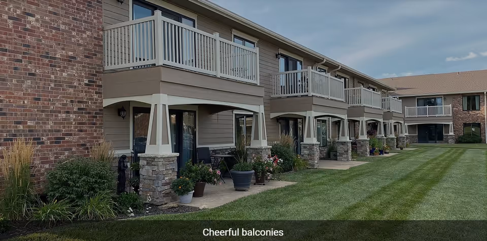 Exterior view of a senior living facility showing a row of ground floor patios and upper floor balconies with white railings. The building has a combination of brick and beige siding with stone pillars supporting the balconies. There are potted plants and outdoor chairs on the patios, and a well-maintained green lawn in front.