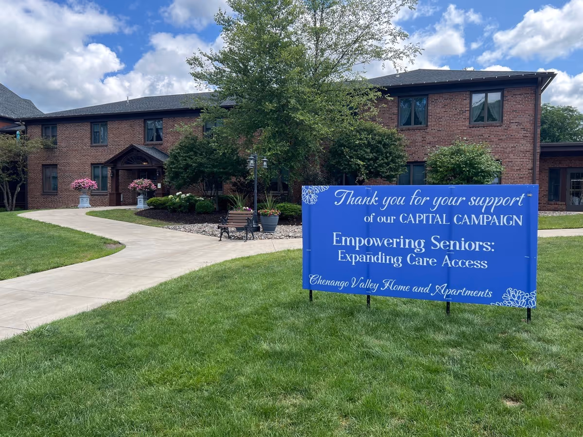 Exterior view of a two-story brick building with a paved walkway leading to the entrance. There are green lawns, trees, bushes, and flower pots near the entrance. A blue sign on the lawn reads: 'Thank you for your support of our CAPITAL CAMPAIGN Empowering Seniors: Expanding Care Access Chenango Valley Home and Apartments.' The sky is partly cloudy.