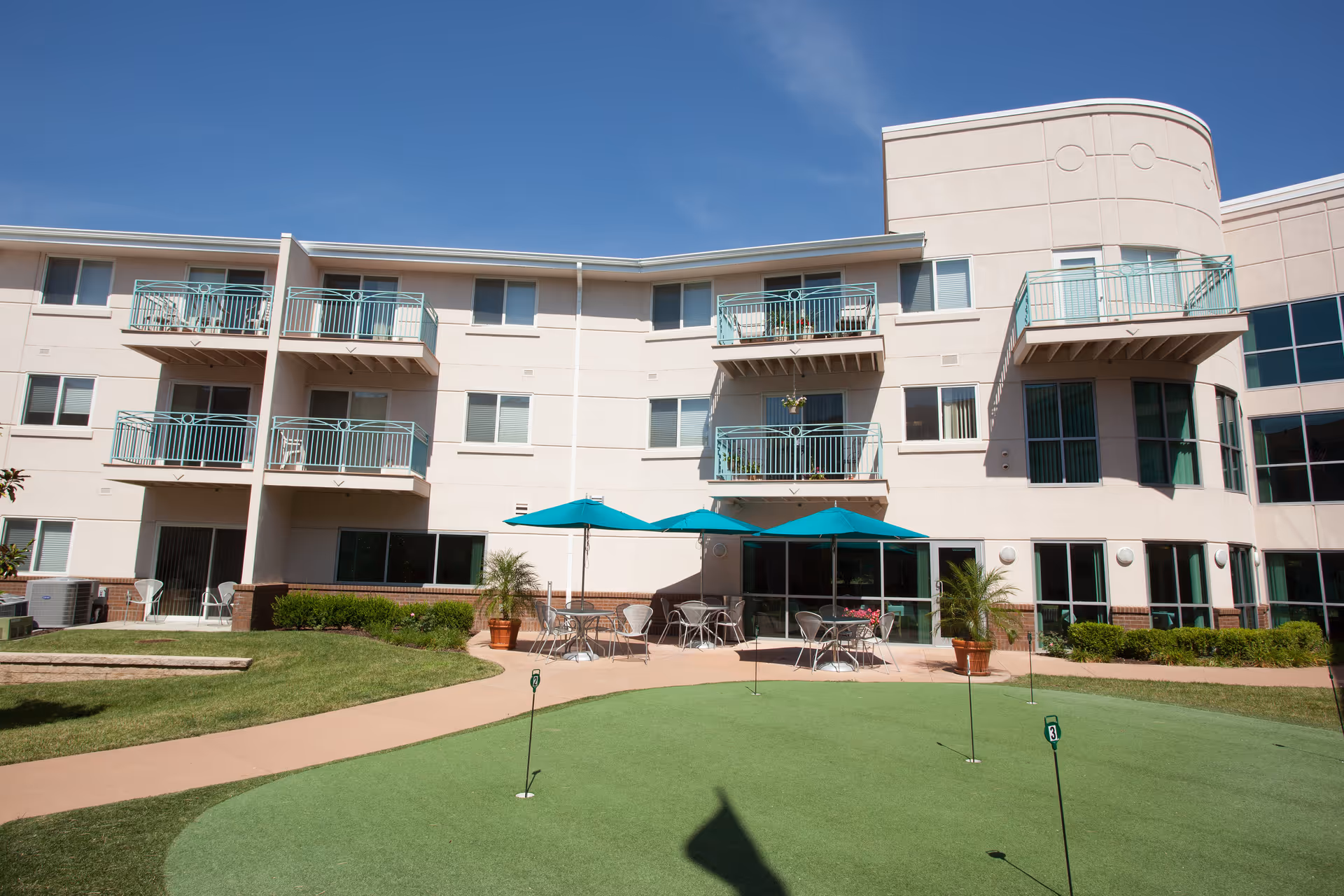 Outdoor view of a senior living facility with a putting green in the foreground, patio tables with teal umbrellas, and a three-story building with balconies and large windows under a clear blue sky.