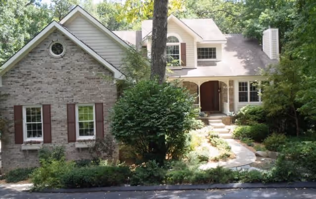 Front exterior view of a two-story brick house with white trim, brown shutters, and a sloped roof surrounded by trees and greenery. A curved walkway leads to the front door, partially shaded by a large tree and bushes.