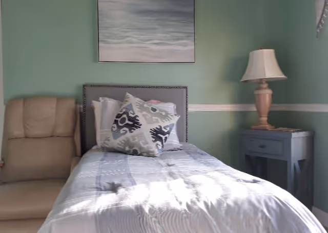A small bedroom with a single bed covered with a light-colored bedspread and decorative pillows. To the left of the bed is a beige armchair, and to the right is a blue nightstand with a beige lamp on top. The walls are painted green with a white chair rail, and a painting of a cloudy sky hangs above the bed.