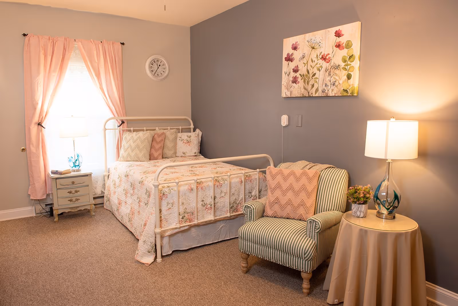 A cozy bedroom with a white metal bed frame adorned with floral bedding and multiple pillows. Next to the bed is a small nightstand with a lamp and some books. A window with pink curtains allows natural light into the room. On the right side, there is a striped armchair with a pink chevron-patterned pillow, next to a round table covered with a beige cloth, holding a lamp and a small potted plant. A floral painting hangs on the gray wall above the armchair.