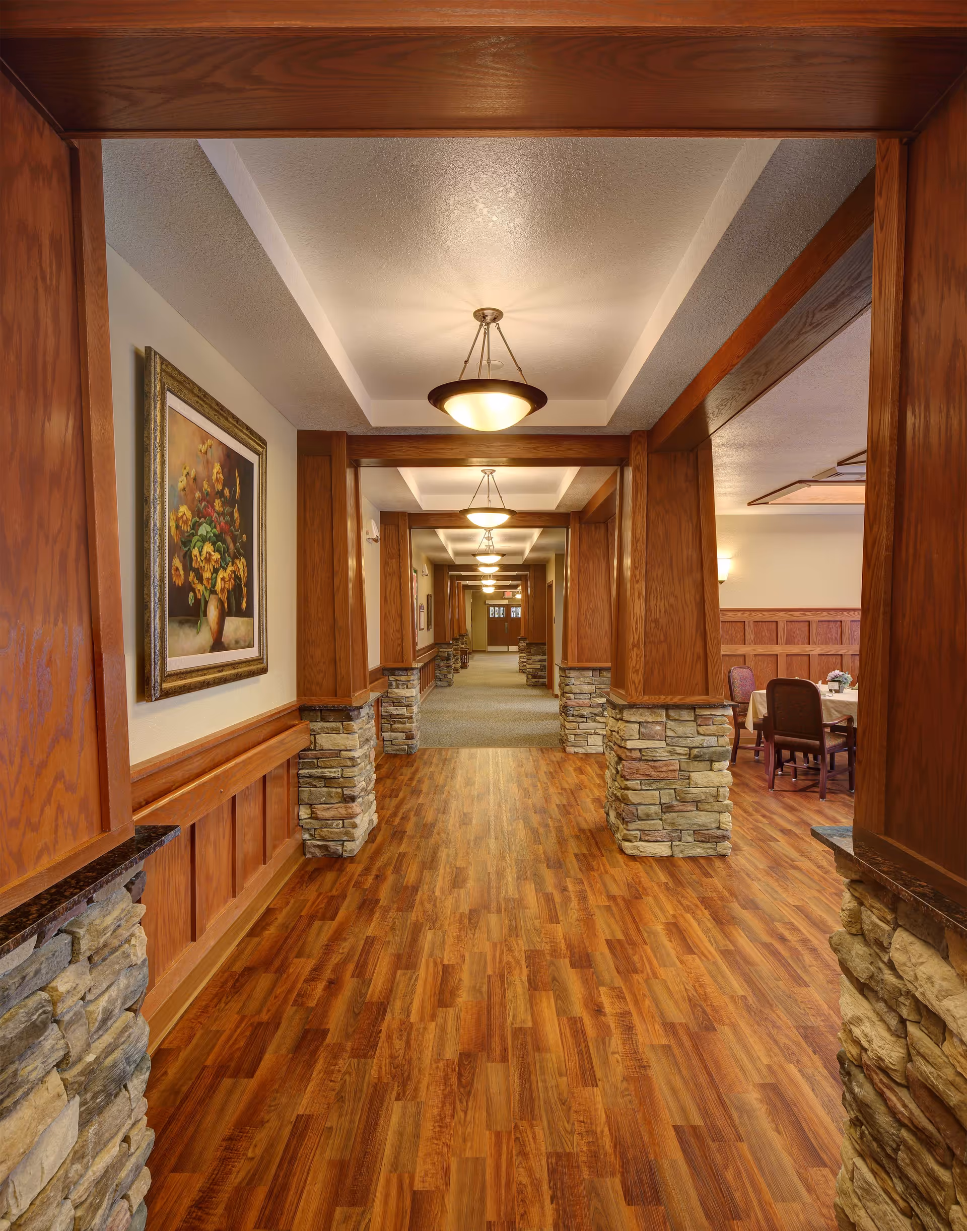 A long hallway in a senior living facility with wooden flooring and stone pillars supporting wooden beams. The ceiling has recessed lighting fixtures, and there is a framed painting of flowers on the left wall. To the right, there is a dining area with tables and chairs.