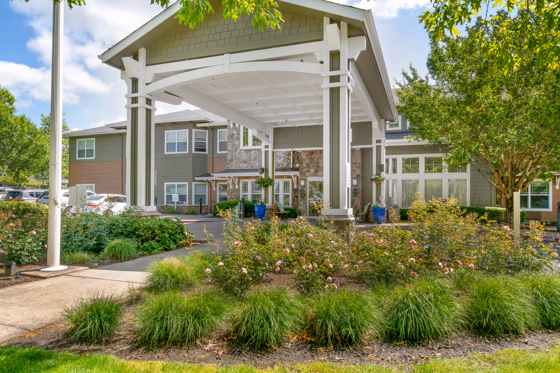 Entrance of a senior living facility with a covered drop-off area supported by white pillars. The building exterior features green and brown siding with stone accents. There are landscaped bushes and flowers in front, along with a sidewalk and a tree to the right. Several parked cars are visible in the background under a partly cloudy sky.
