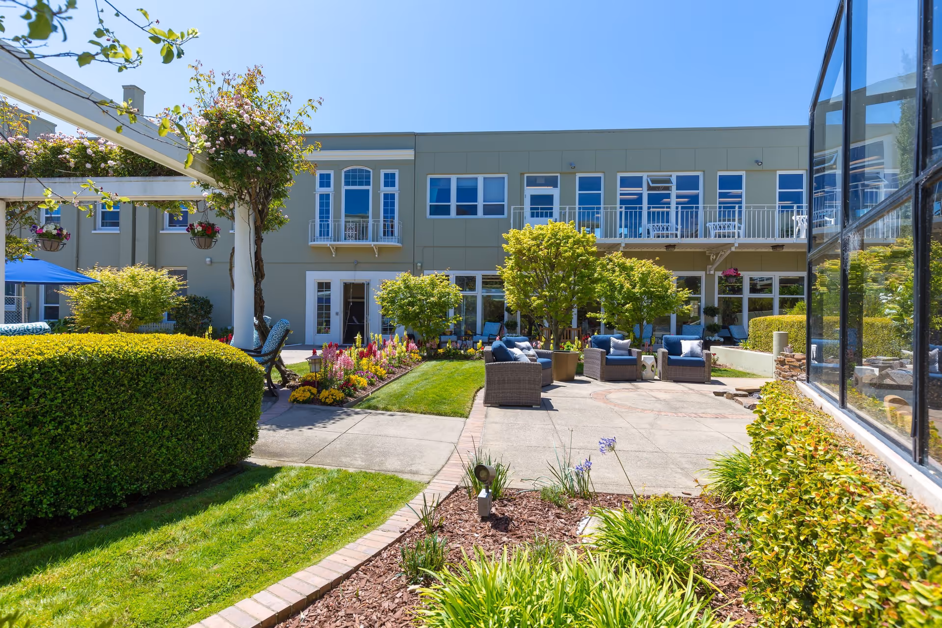 Outdoor patio area at Sagebrook Senior Living with comfortable seating arrangements, surrounded by well-maintained greenery, flowering plants, and trees. The building in the background has large windows and balconies overlooking the garden. The sky is clear and blue.