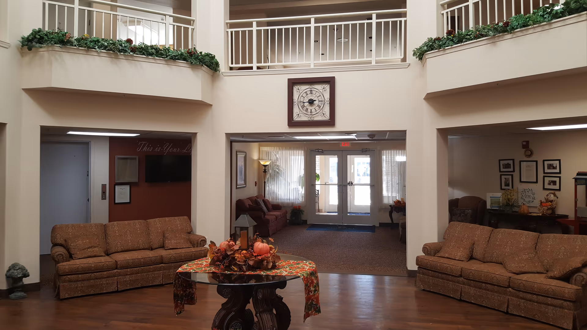 Interior view of a senior living facility lobby with two patterned sofas facing each other, a round wooden table with a fall-themed centerpiece in the middle, and a large clock mounted on the wall above an open hallway leading to glass double doors. The upper level has white railings with green plants along the edges.