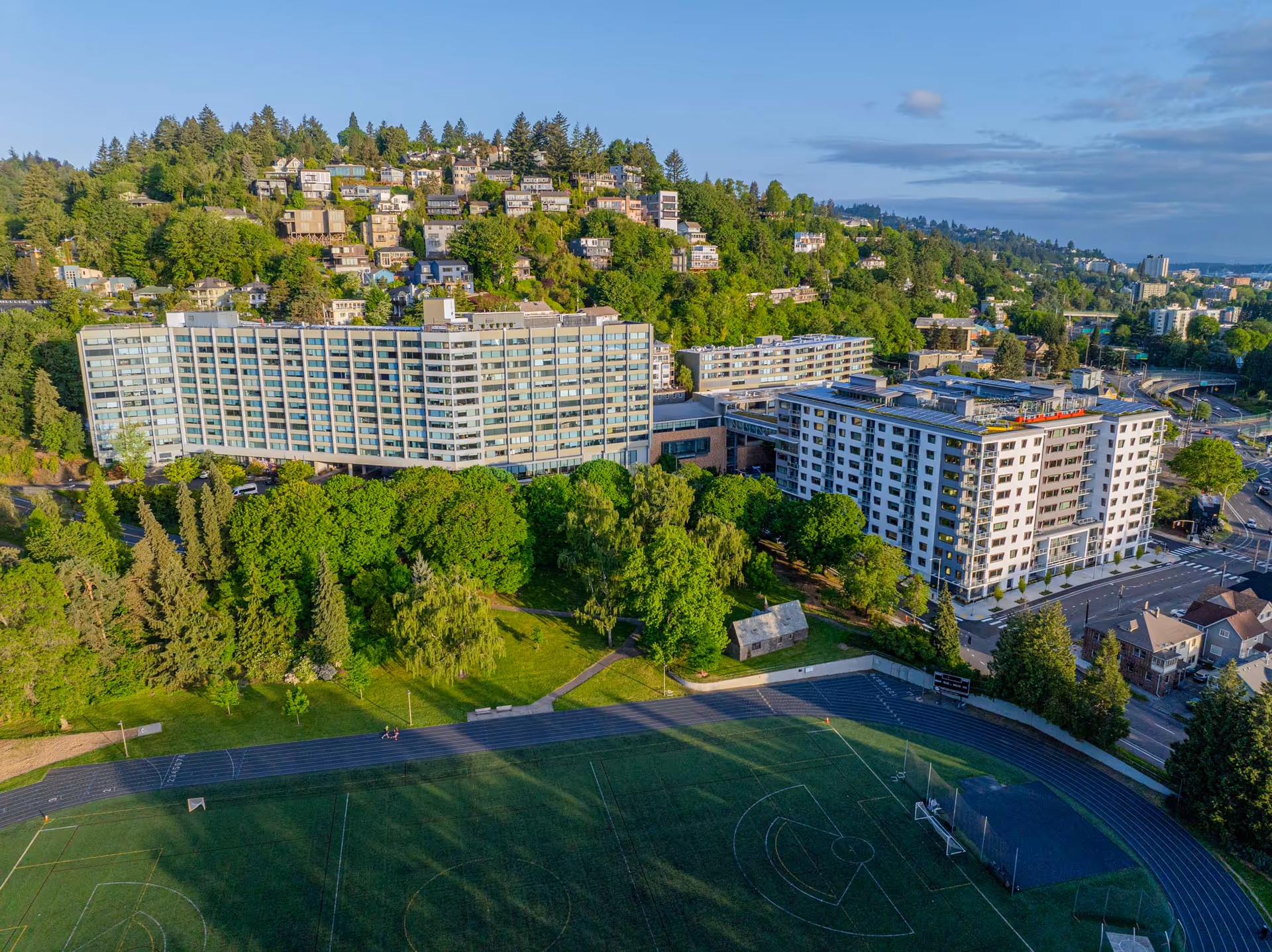 Aerial view of Terwilliger Plaza Retirement Community, a large multi-story building surrounded by lush green trees and residential houses on a hillside. In the foreground, there is a sports field with a running track and soccer goals. The sky is partly cloudy with blue patches.