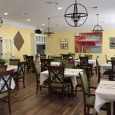 A dining room with multiple tables covered with white tablecloths and green cushioned wooden chairs. The room has yellow walls adorned with colorful abstract paintings and decorative wall hangings. Modern spherical pendant lights hang from the ceiling, and the floor is wooden.