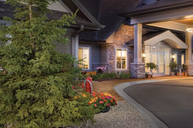 Exterior view of The Moments Memory Care facility entrance at dusk, featuring a covered driveway, stone and siding facade, illuminated windows, and landscaped greenery including a large pine tree and colorful flowers.