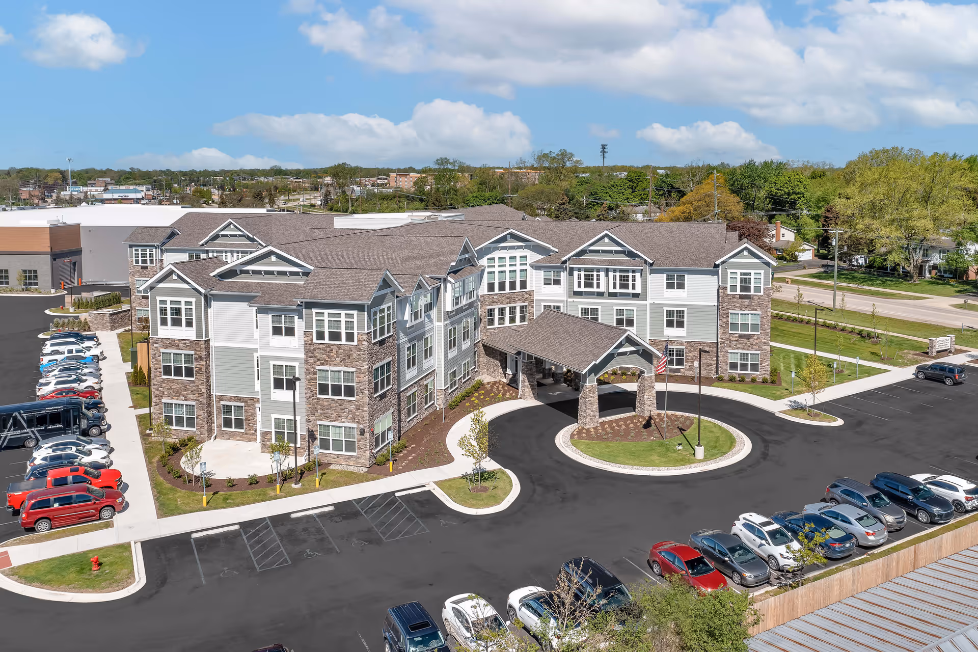 Aerial view of StoryPoint Farmington Hills, a large multi-story senior living facility with a covered entrance and surrounding parking lot filled with cars. The building features a combination of stone and light-colored siding with multiple windows. The area around the building is landscaped with small trees and grass, and a clear blue sky with some clouds is visible above.