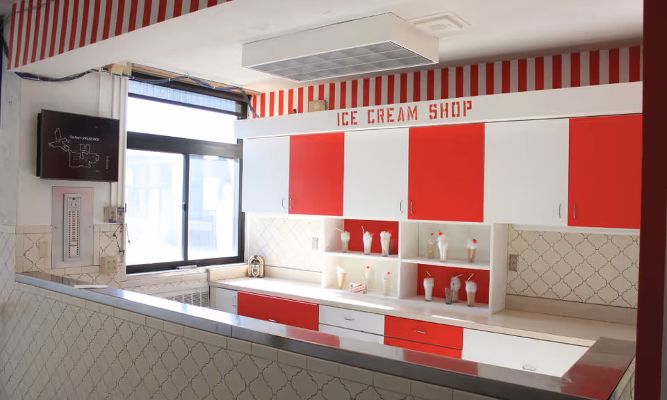 Interior view of an ice cream shop counter with red and white cabinets and a sign reading 'ICE CREAM SHOP' above. The counter has several plastic ice cream sundae models displayed on shelves. There is a window on the left side letting in natural light, and the walls have a patterned tile design with red and white striped wallpaper near the ceiling.